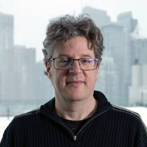 Bart Simon is standing in a black open-collared jumper, with black narrow-rimmed glasses with the view from the 11th floor of the EV building out of focus behind hm.